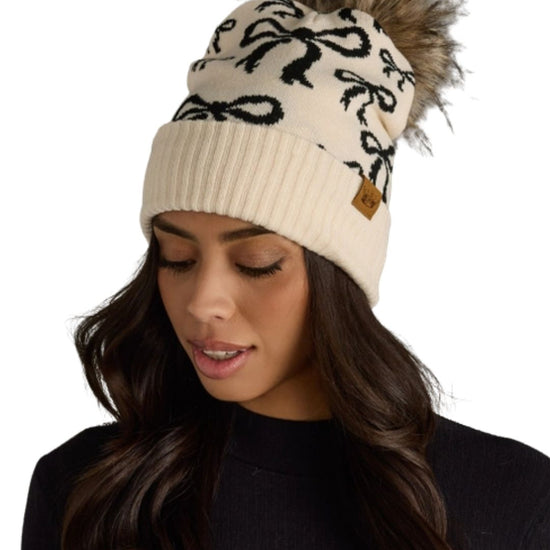 Beige beanie with black bows and fur pom-pom worn by a model on a white background - equestrian fashion outfitters