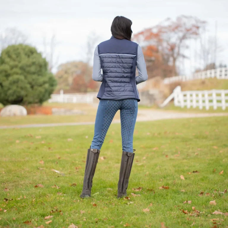 Person wearing a blue EQuinavia Heidi insulated vest, gray breeches, and black equestrian riding boots standing in a grassy area