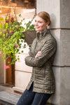 Woman in a green quilted jacket leaning against a stone wall with plants in the background