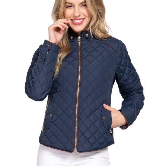 A woman wearing a navy blue diamond-quilted jacket with brown trim and a gold zipper, paired with white pants, against a plain white background equestrian fashion outfitters