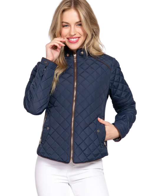 A woman wearing a navy blue diamond-quilted jacket with brown trim and a gold zipper, paired with white pants, against a plain white background equestrian fashion outfitters