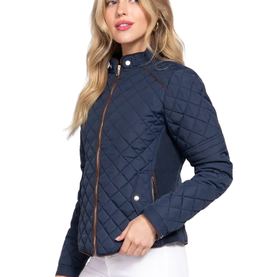 A woman modeling a navy blue diamond-quilted jacket with brown trim and silver snap buttons, paired with white pants, against a plain white background equestrian fashion outfitters