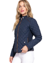 A woman modeling a navy blue diamond-quilted jacket with brown trim and silver snap buttons, paired with white pants, against a plain white background equestrian fashion outfitters