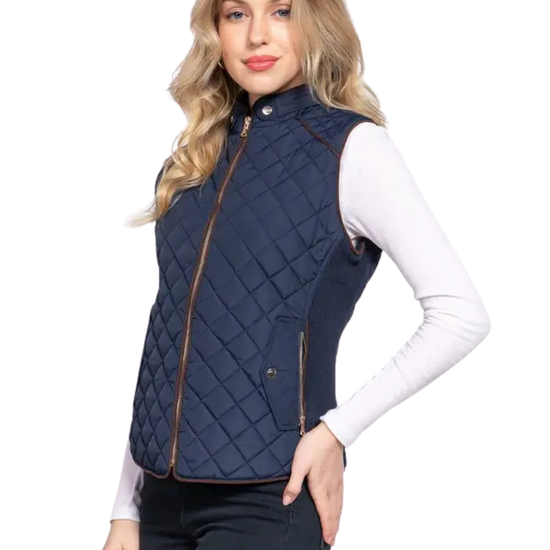 A woman modeling a navy blue quilted sleeveless vest with brown trim over a white long-sleeve top and dark pants, against a plain white background. equestrian fashion outfitters