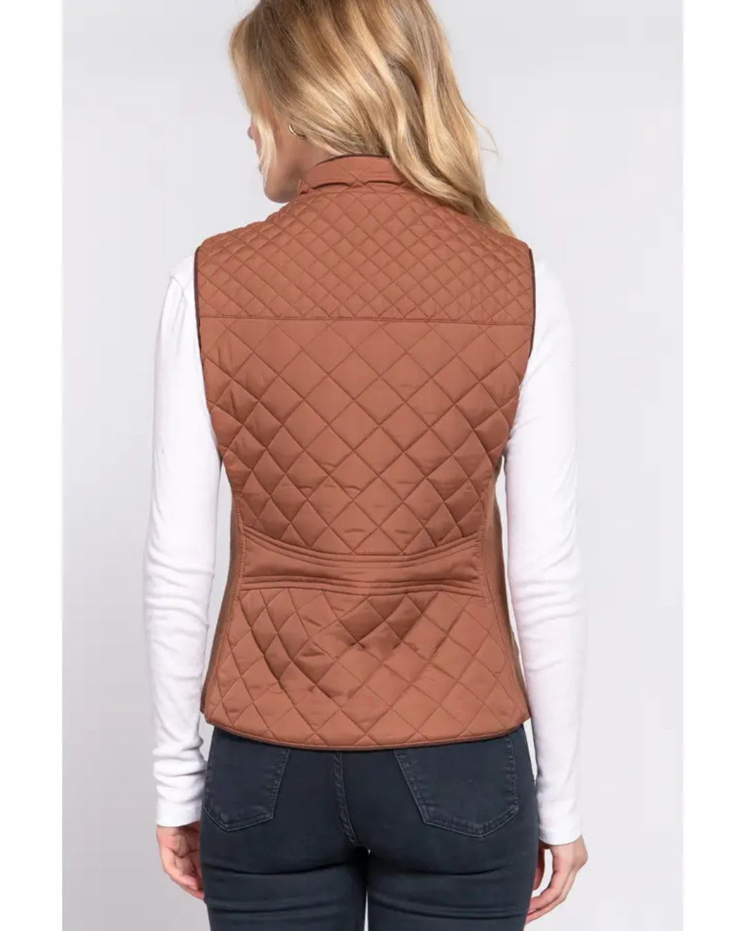 A woman shown from the back wearing a quilted sleeveless vest in a warm rust brown camel color over a white long-sleeve shirt and dark jeans, equestrian fashion outfitters.