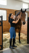 Woman wearing a pair of blue riding pants with black accents petting a horse equestrian fashion outfitters
