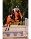Glossy black riding helmet with wide brim mips girl on brown horse jumping a jump with red breeches. equestrian fashion outfitters