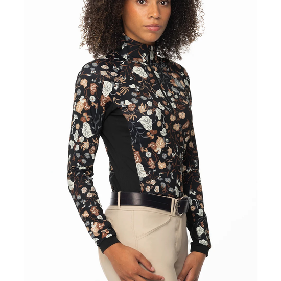 A woman modeling a long-sleeve 1/4 zip equestrian shirt with a black base and a detailed floral and vine pattern in beige, brown, and white, with black side panels paired with beige riding pants equestrian fashion outfitters