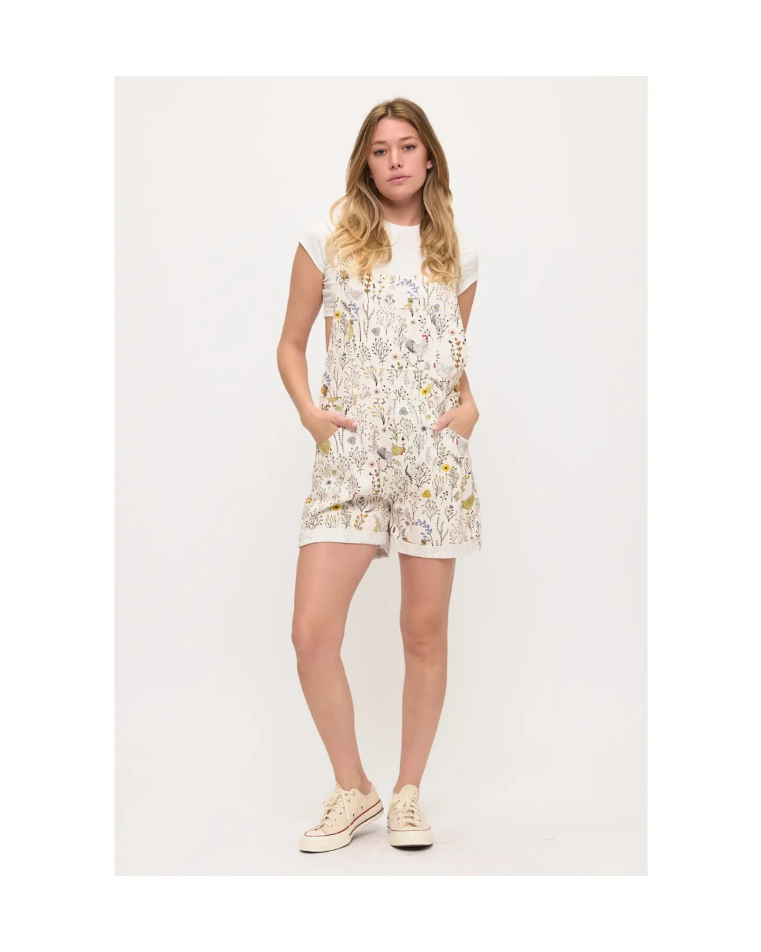 A woman modeling overalls or shortalls with roosters and chickens and flowers on a white background with sneakers equestrian fashion outfitters