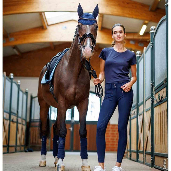 Woman in equestrian attire standing next to a horse in a stable. schockemohle tulip equestrian fashion outfitters