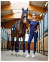 Woman in equestrian attire standing next to a horse in a stable. schockemohle tulip equestrian fashion outfitters