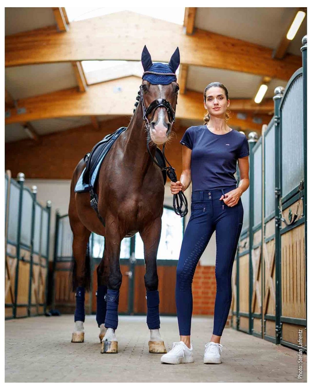Woman in equestrian attire standing next to a horse in a stable. schockemohle tulip equestrian fashion outfitters