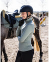 woman standing wearing a helmet and HKM Supersoft II Functional 1/4 Zip Shirt in sage green with equestrian background