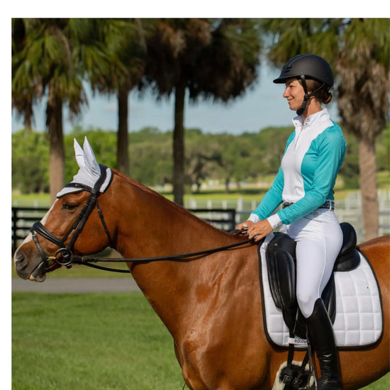 Woman riding a horse in an equestrian setting with trees in the background with a white saddle pad