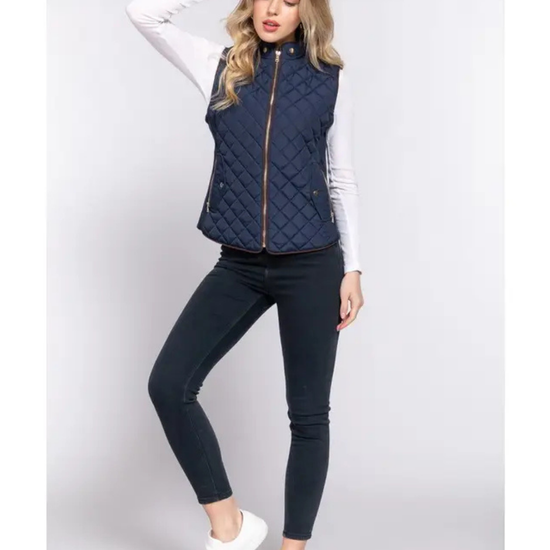A woman modeling a navy blue quilted sleeveless quilted vest over a white long-sleeve shirt, paired with dark skinny jeans and white sneakers, equestrian fashion outfitters