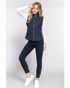 A woman modeling a navy blue quilted sleeveless quilted vest over a white long-sleeve shirt, paired with dark skinny jeans and white sneakers, equestrian fashion outfitters