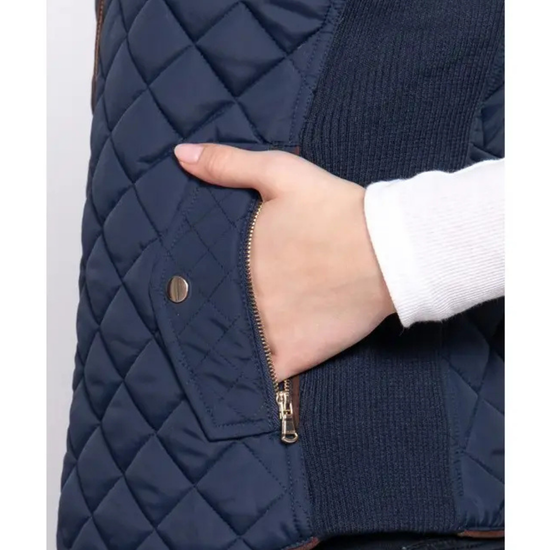 Close-up of a navy blue quilted vest with a hand in a white sleeve, showing a diagonal zip pocket and a gold snap button equestrian fashion outfitters