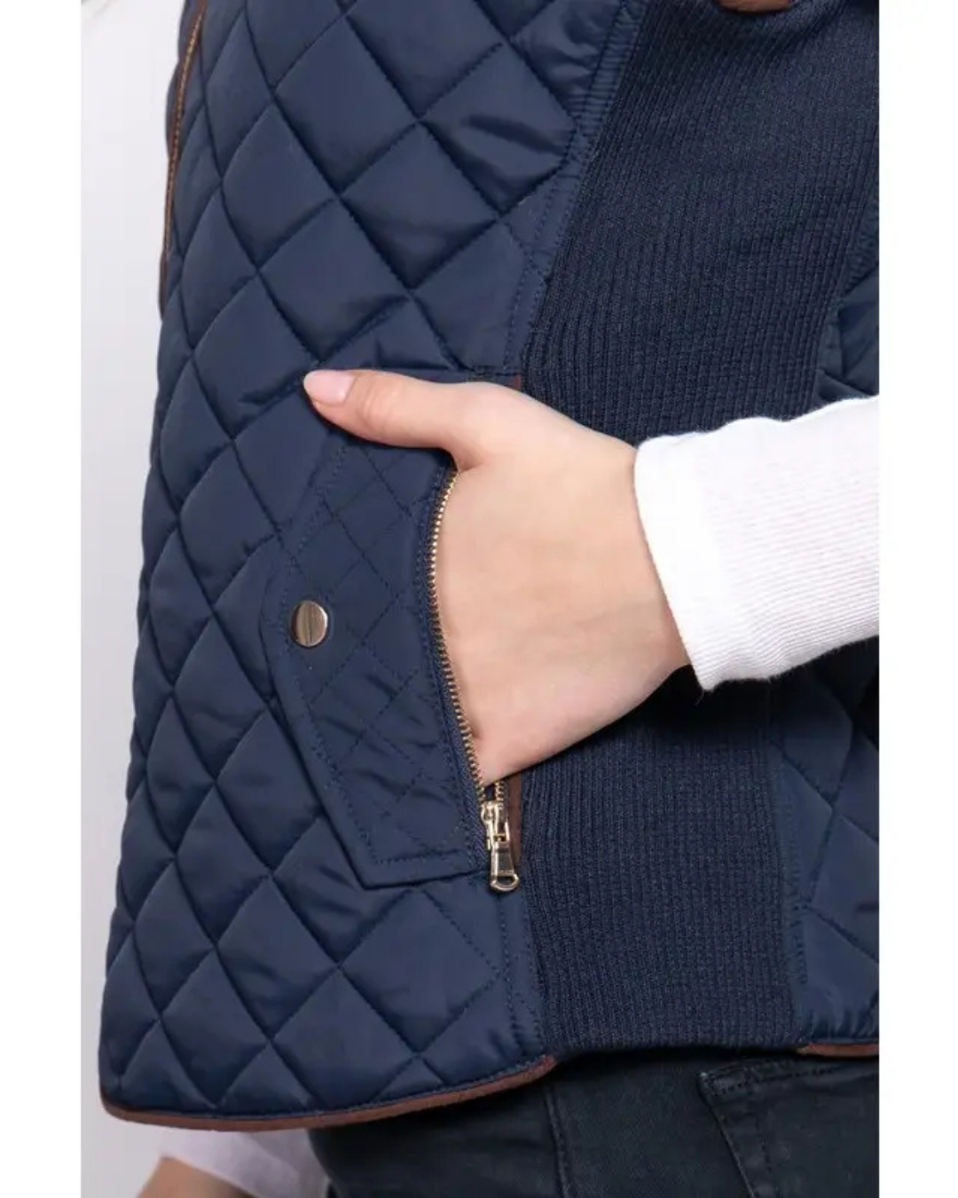Close-up of a navy blue quilted vest with a hand in a white sleeve, showing a diagonal zip pocket and a gold snap button equestrian fashion outfitters