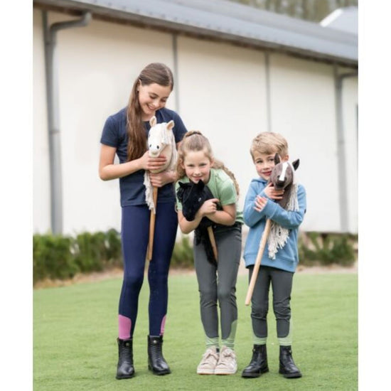 Three children and a woman holding small stick horses on a grassy field. equestrian fashion outfitters