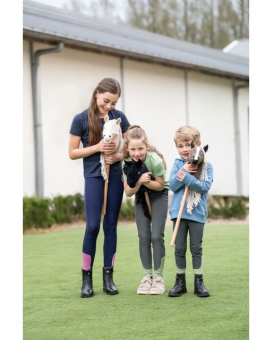 Three children and a woman holding small stick horses on a grassy field. equestrian fashion outfitters