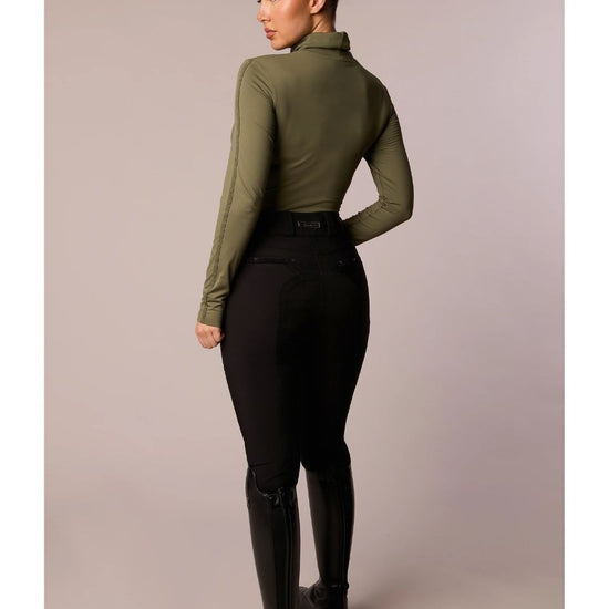 Person wearing an olive green long-sleeve top and black riding pants on a plain background