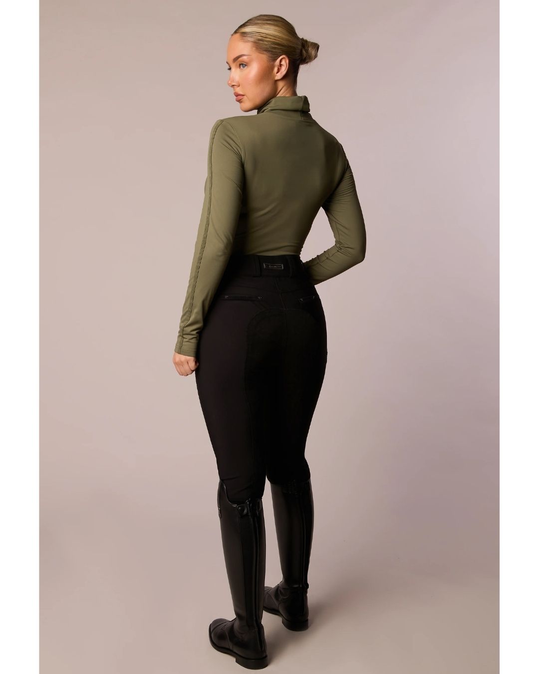 Person wearing an olive green long-sleeve top and black riding pants on a plain background