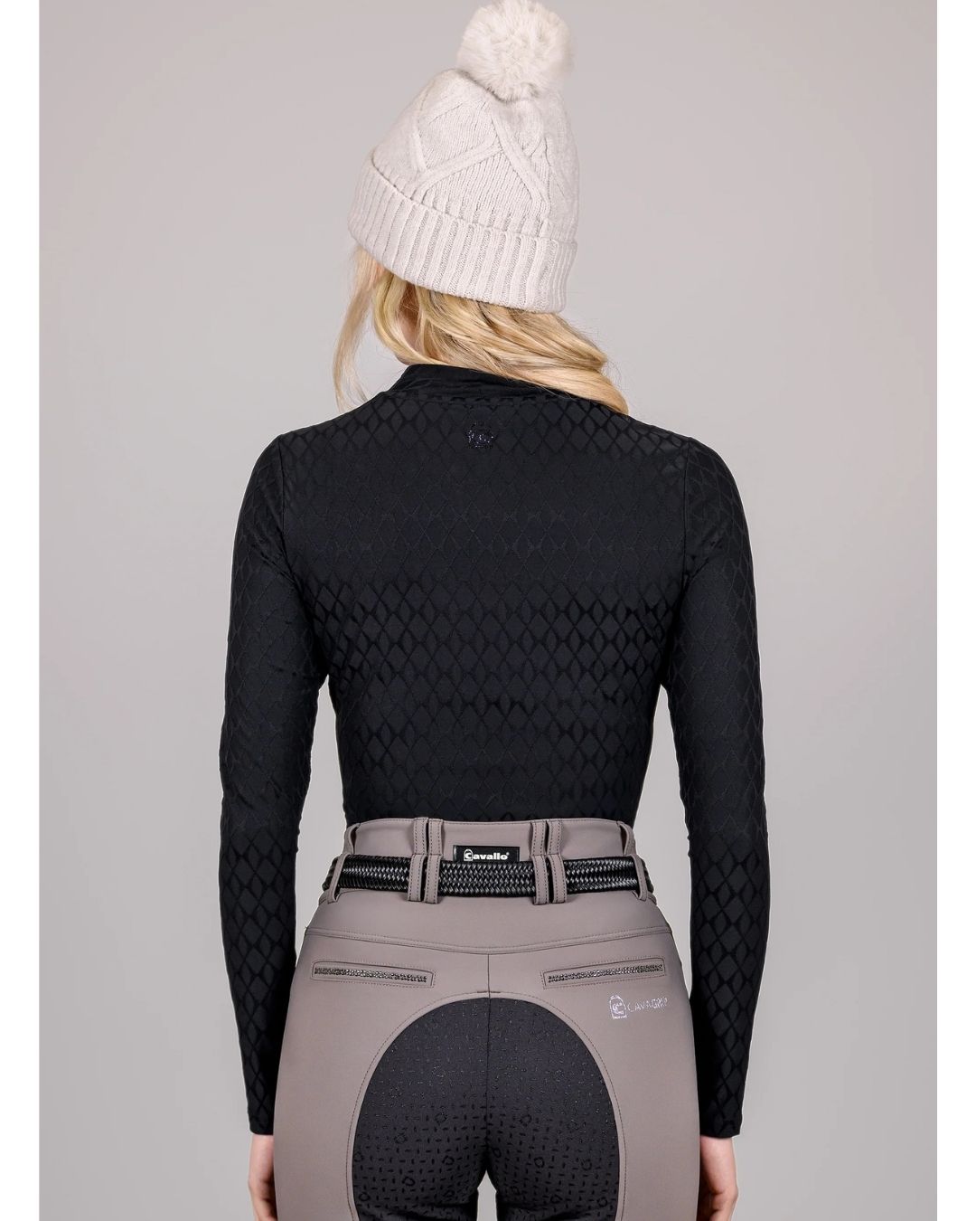Person wearing a black long-sleeve top and beige riding pants with a white knit cap on a gray background