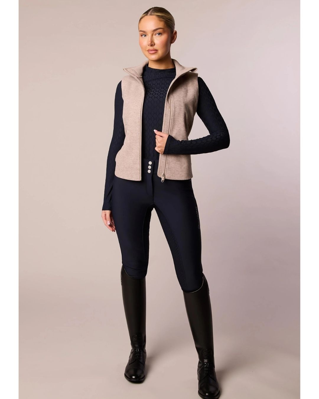 Person wearing equestrian attire with a beige vest and black riding pants on a plain background