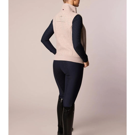 Person wearing a beige vest, navy long-sleeve shirt, dark pants, and black riding boots on a plain background