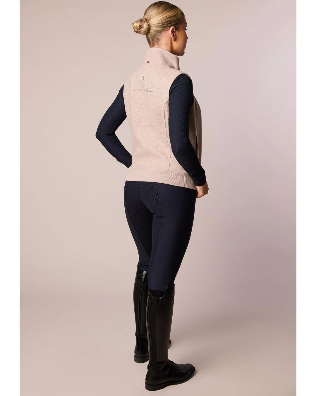 Person wearing a beige vest, navy long-sleeve shirt, dark pants, and black riding boots on a plain background