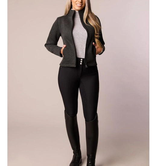 Woman wearing a gray black jacket, gray top, black riding pants, and black boots on a beige background
