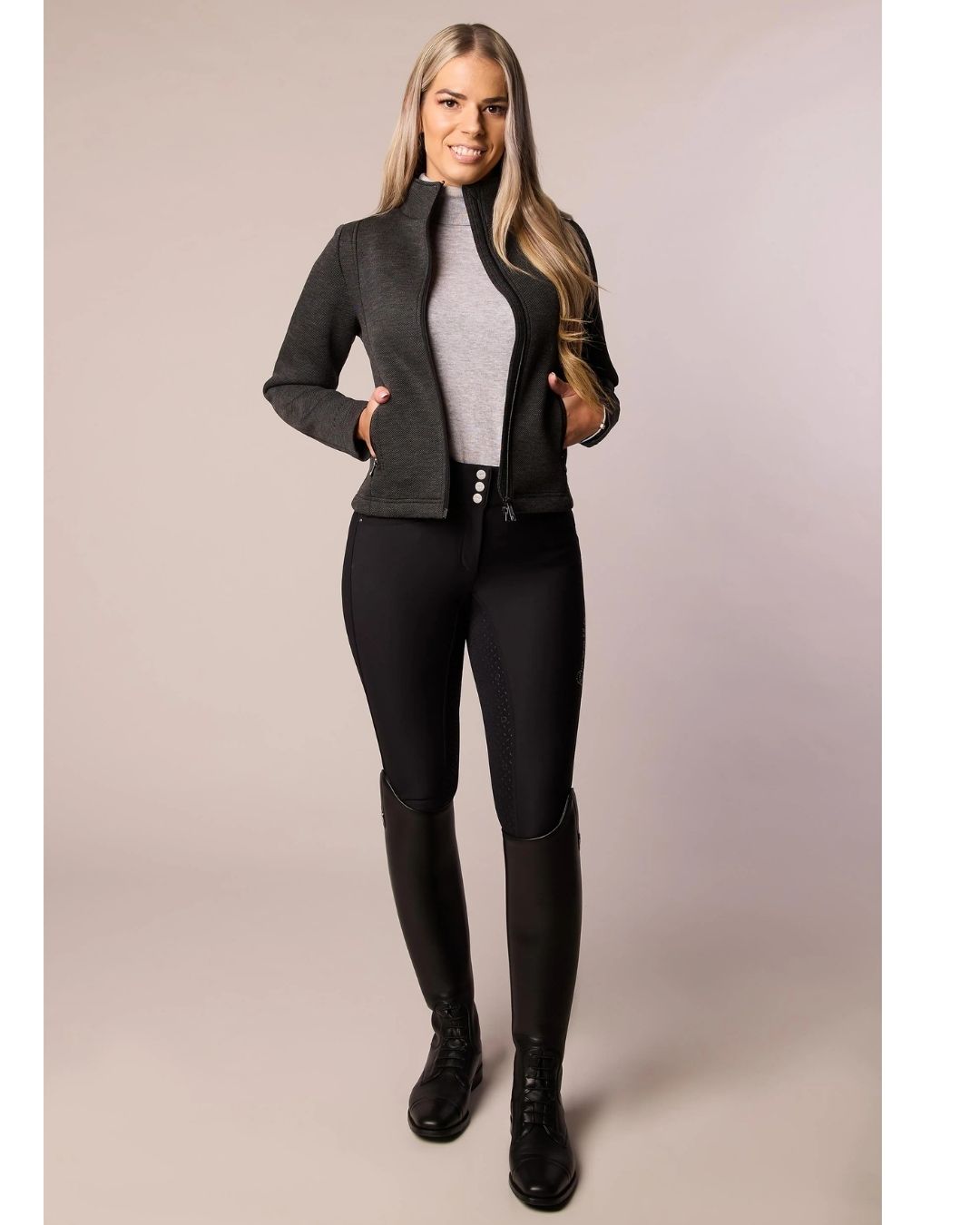 Woman wearing a gray black jacket, gray top, black riding pants, and black boots on a beige background