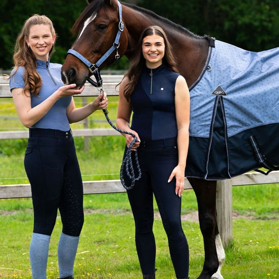 Two women in equestrian attire standing next to a horse with a blue blanket in an outdoor setting qhp aubree shirt and breeches equestrian fashion outfitters