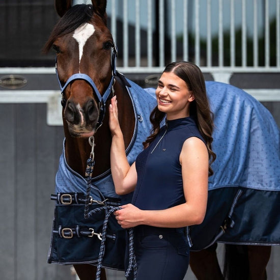 Woman in equestrian attire standing next to a horse wearing a blue blanket. equestrian fashion outfitters