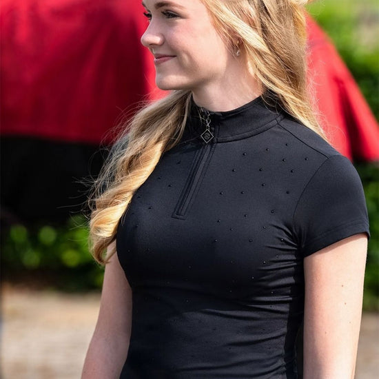 Woman wearing a black equestrian shirt with a horse in the background equestrian fashion outfitters