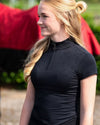 Woman wearing a black equestrian shirt with a horse in the background equestrian fashion outfitters