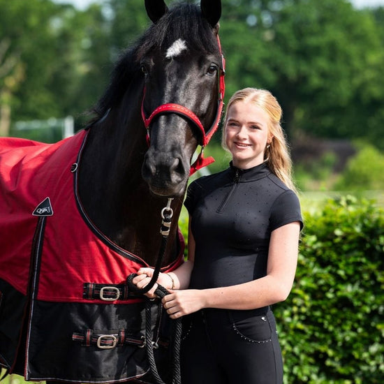 Woman in equestrian attire standing next to a black horse wearing a red rug in an outdoor setting. equestrian fashion outfitters