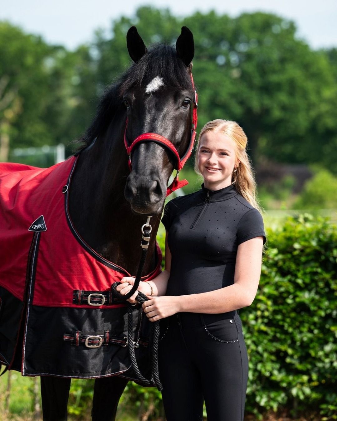 Woman in equestrian attire standing next to a black horse wearing a red rug in an outdoor setting. equestrian fashion outfitters