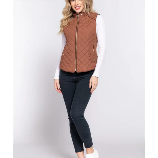 A woman modeling a rust-brown camel quilted sleeveless zip-front vest over a white long-sleeve top, black skinny pants, and white slip-on shoes, equestrian fashion outfitters