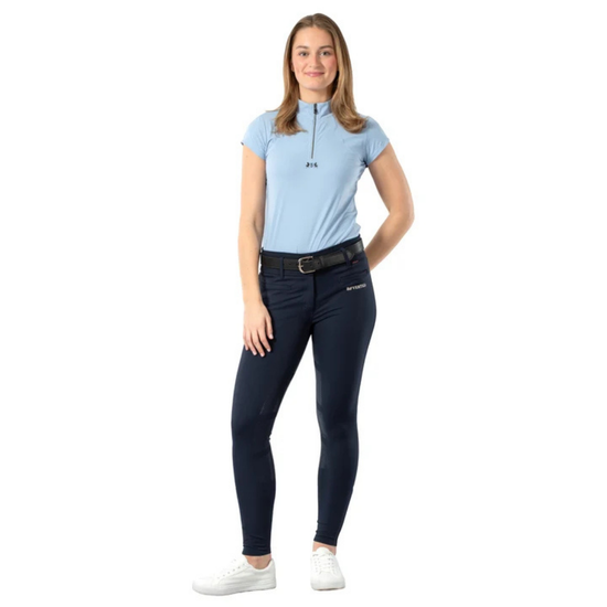 A woman modeling a light blue short-sleeve polo shirt with a half-zip neckline, paired with dark navy equestrian riding breeches and white sneakers, set against a plain white background. equestrian fashion outfitters