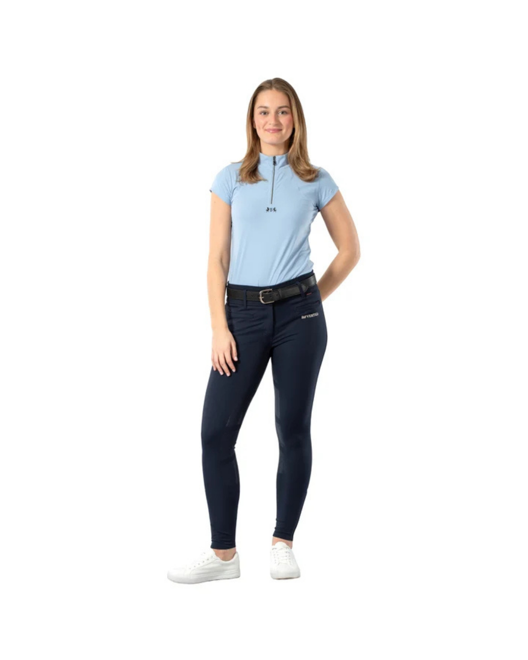 A woman modeling a light blue short-sleeve polo shirt with a half-zip neckline, paired with dark navy equestrian riding breeches and white sneakers, set against a plain white background. equestrian fashion outfitters