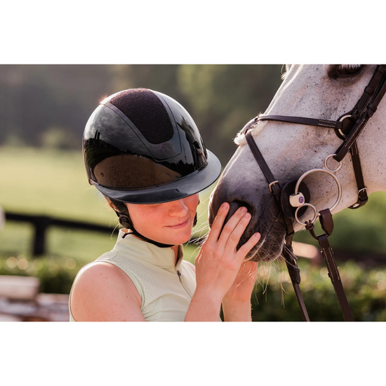 A woman in a light green sleeveless top wears a glossy black equestrian riding helmet with a diamond sparkle top panel and gently touches a white horse's face in an outdoor setting equestrian fashion outfitters