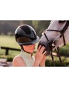 A woman in a light green sleeveless top wears a glossy black equestrian riding helmet with a diamond sparkle top panel and gently touches a white horse's face in an outdoor setting equestrian fashion outfitters