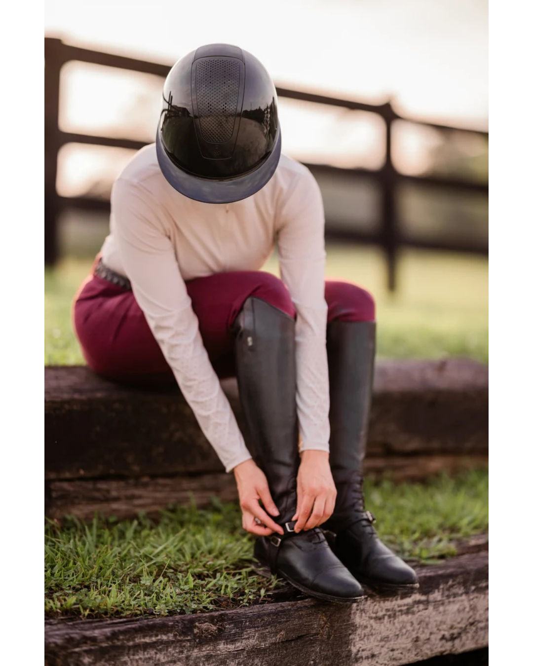 Girl wearing a black glossy  shiny helmet with burgundy breeches tying up her boots equestrian fashion outfitters