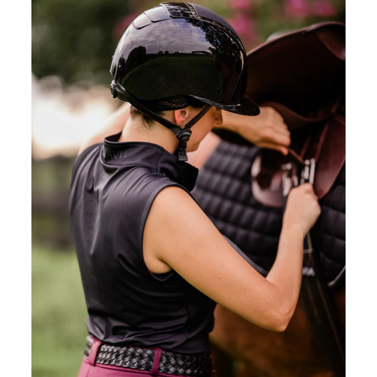girl with a black shiny glossy helmet with vents and a shimmer top panel with black straps doing up a horses girth wearing a black sleeveless shirt equestrian fashion outfitters