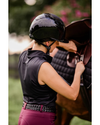 girl with a black shiny glossy helmet with vents and a shimmer top panel with black straps doing up a horses girth wearing a black sleeveless shirt equestrian fashion outfitters