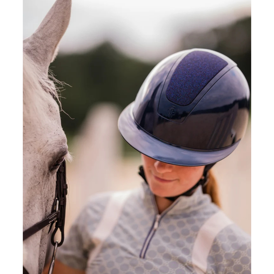 A close-up lifestyle shot of a person wearing a dark navy blue glittery shimmer equestrian riding helmet and a grey patterned riding shirt, standing next to a white horse in an outdoor setting equestrian fashion outfitters