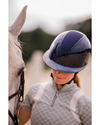 A close-up lifestyle shot of a person wearing a dark navy blue glittery shimmer equestrian riding helmet and a grey patterned riding shirt, standing next to a white horse in an outdoor setting equestrian fashion outfitters