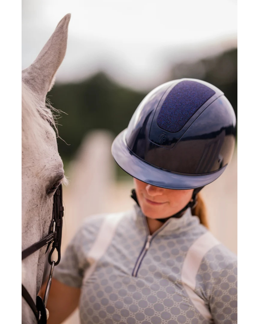 A close-up lifestyle shot of a person wearing a dark navy blue glittery shimmer equestrian riding helmet and a grey patterned riding shirt, standing next to a white horse in an outdoor setting equestrian fashion outfitters