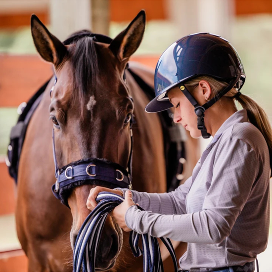 girl with navy blue shiny glossy helmet with black harness straps and yellow mips logo fixing a lunge line on a horse equestrian fashion outfitters